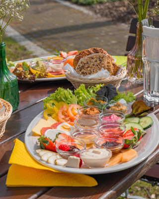 Tisch im Außenbereich. Auf dem Tisch steht ein kleines Körbchen mit Brötchen und ein großer weißer Teller mit Aufstrichen und Aufschnitt